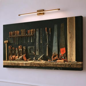 May include: A rectangular wall art print depicting a dark workbench surface with an array of woodworking tools including chisels, hand planes, metal rulers, and two hand saws, all illuminated by a slim, horizontal gold-toned LED light fixture.