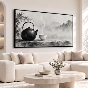 Puede incluir: Un cuadro de pared en blanco y negro que muestra una tetera de hierro oscuro y una pequeña taza humeante sobre un saliente rocoso. El fondo representa un paisaje brumoso con montañas y árboles tenues, enmarcado en negro.