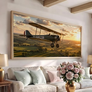 May include: A rectangular wall art print in a light wood frame depicting a vintage biplane flying over a green countryside at sunset. The dark-colored aircraft features a circular insignia on its side and is captured mid-flight against a golden sky.
