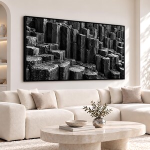 May include: A large, rectangular, black and white wall art print featuring a detailed landscape of vertical basalt rock columns. The artwork is encased in a thin black frame and is displayed horizontally above a light-colored sofa.
