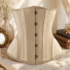 May include: A beige corset featuring a front busk closure with five metal clasps. The garment is constructed with vertical panels of solid beige fabric alternating with lighter beige textured panels that display a subtle, tone-on-tone floral pattern.