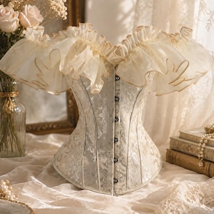 May include: A cream-colored corset featuring a subtle floral brocade pattern, a front busk closure with metal clasps, and an off-the-shoulder neckline adorned with voluminous, layered organza ruffles that have a delicate, light brown trim.