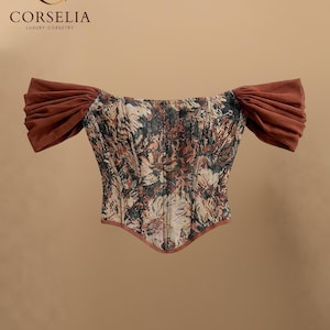 May include: A corset top featuring a floral tapestry pattern in shades of brown, black, and gold. The garment has off-the-shoulder sleeves made of solid brown fabric and a pointed hemline. The text CORSELIA LUXURY CORSETRY is visible.