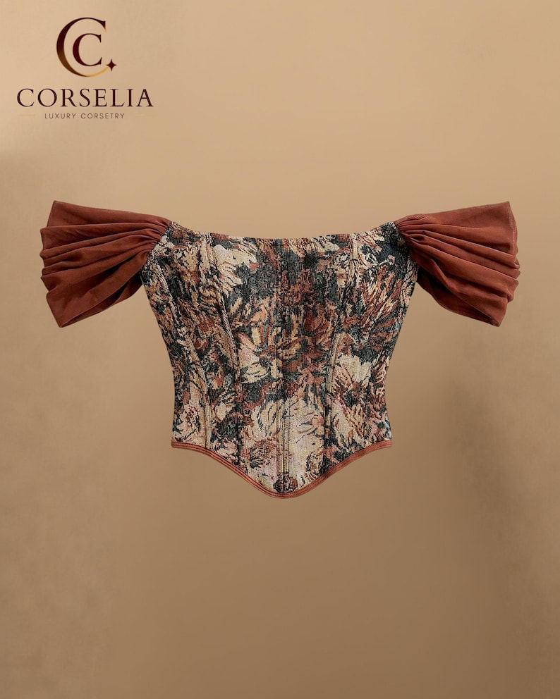 May include: A corset top featuring a floral tapestry pattern in shades of brown, black, and gold. The garment has off-the-shoulder sleeves made of solid brown fabric and a pointed hemline. The text CORSELIA LUXURY CORSETRY is visible.
