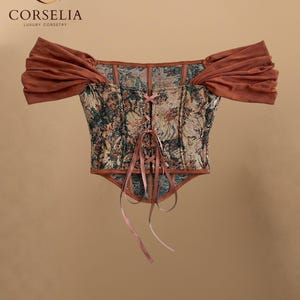 May include: A corset top featuring a multi-colored floral tapestry pattern with a front lace-up closure using thin ribbons. The garment includes off-the-shoulder sleeves made of solid rust-colored fabric and a pointed hemline at the bottom.