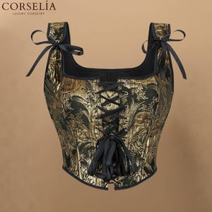 May include: A black and gold corset top featuring a square neckline, wide shoulder straps with black ribbon ties, and a front lace-up closure with black ribbons. The fabric displays a metallic gold botanical pattern on a black background.