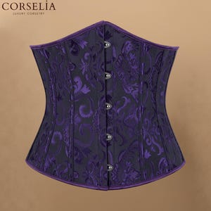 May include: A dark purple corset featuring a black damask pattern, a front busk closure with five silver-toned metal clasps, and purple piping along the top and bottom edges. The text CORSELIA LUXURY CORSETRY is visible in the upper left corner.