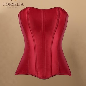 Puede incluir: Un corsé rojo oscuro sin tirantes con un acabado de satén suave y paneles verticales. La esquina superior izquierda muestra el texto CORSELIA LUXURY CORSETRY con un logotipo de C estilizado. La prenda tiene un escote corazón y un corpiño estructurado.