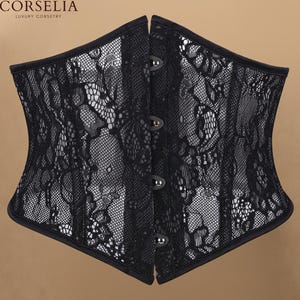May include: A black corset featuring a floral lace overlay on a mesh base. The garment has a front closure with four silver-toned metal busk clasps. The text CORSELIA LUXURY CORSETRY is visible in the upper left corner of the image.