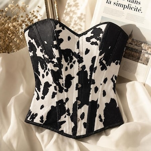 May include: A strapless corset top featuring a black and white cow print pattern. The garment has a sweetheart neckline, vertical paneling, and a front closure with small silver-toned metal hooks. The fabric has a structured, fitted appearance.