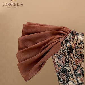 May include: A corset featuring a brown, black, and tan floral tapestry pattern with a large, pleated, sheer brown fabric sleeve detail. The top left corner displays the text CORSELIA LUXURY CORSETRY with a stylized C logo above the brand name.