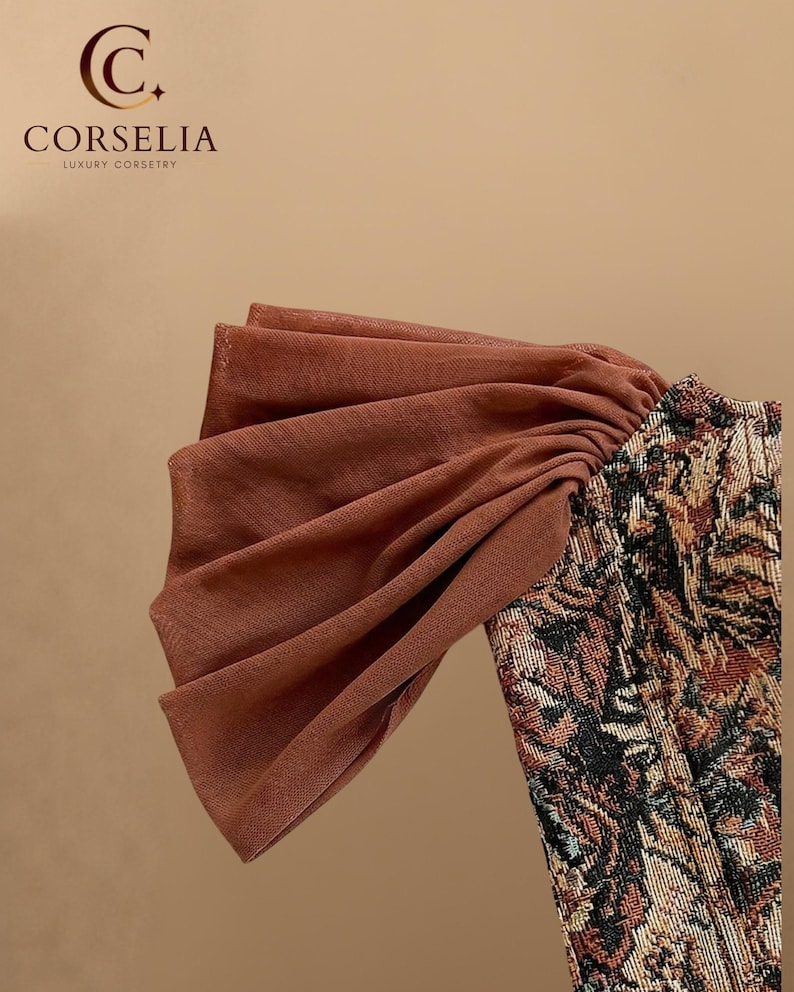 May include: A corset featuring a brown, black, and tan floral tapestry pattern with a large, pleated, sheer brown fabric sleeve detail. The top left corner displays the text CORSELIA LUXURY CORSETRY with a stylized C logo above the brand name.