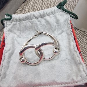 Puede incluir: Una pulsera plateada pulida que presenta dos eslabones circulares entrelazados con extremos redondeados, sobre una bolsa de tela gris claro con cordones verde oscuro. La pulsera tiene un mecanismo de cierre de bisagra visible arriba.