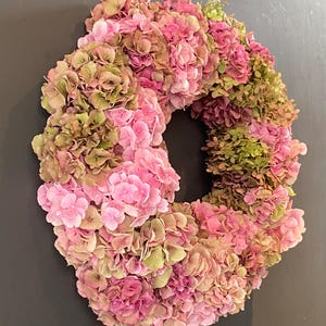 Könnte beinhalten: Ein kreisförmiger Kranz aus getrockneten Hortensienblüten in verschiedenen Schattierungen von Rosa, Magenta und gedämpftem Olivgrün. Die dichte Anordnung der Blütenbüschel erzeugt eine texturierte, runde Form mit einer hohlen Mitte.