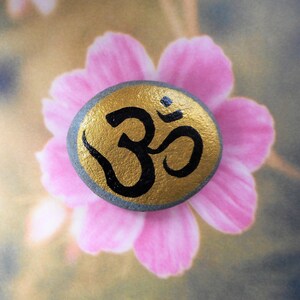 May include: A gray painted rock with a gold Om symbol painted on it. The rock is sitting on a pink flower.