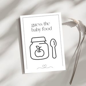 May include: A white rectangular game card featuring the black text "guess the baby food" at the top. Below the text is a black line drawing of a baby food jar with an apple icon on the front and a spoon next to it. A small heart icon is at the bottom.