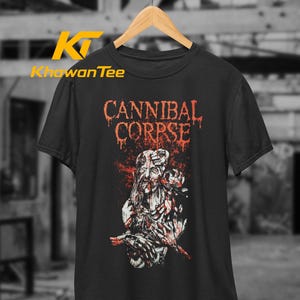 May include: A black short-sleeved t-shirt featuring the text CANNIBAL CORPSE in a dripping red font above a graphic illustration of a decaying figure with exposed bone and red blood splatters, hanging on a wooden hanger against a blurred background.