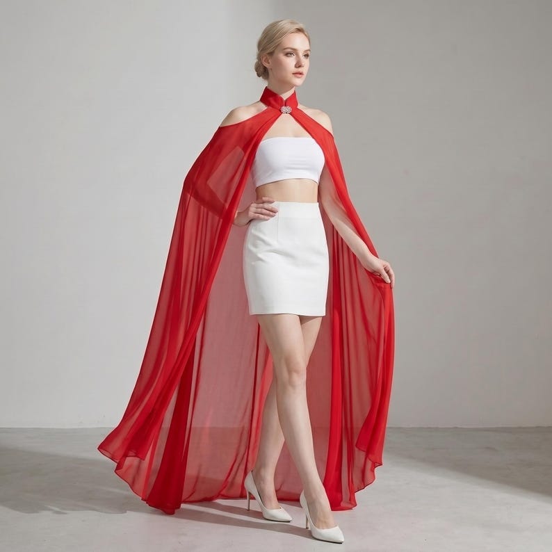May include: A floor-length red chiffon cape with a high collar and a silver-toned clasp, worn over a white strapless crop top and a matching white mini skirt. The cape features open shoulders and a sheer, flowing fabric that drapes to the floor.