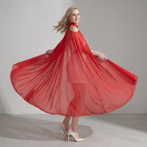 May include: A vibrant red, floor-length dress featuring a sheer, flowing cape overlay that extends from the shoulders. The garment has a high neckline with cold-shoulder cutouts and a fitted bodice, creating a dramatic, wide silhouette in motion.