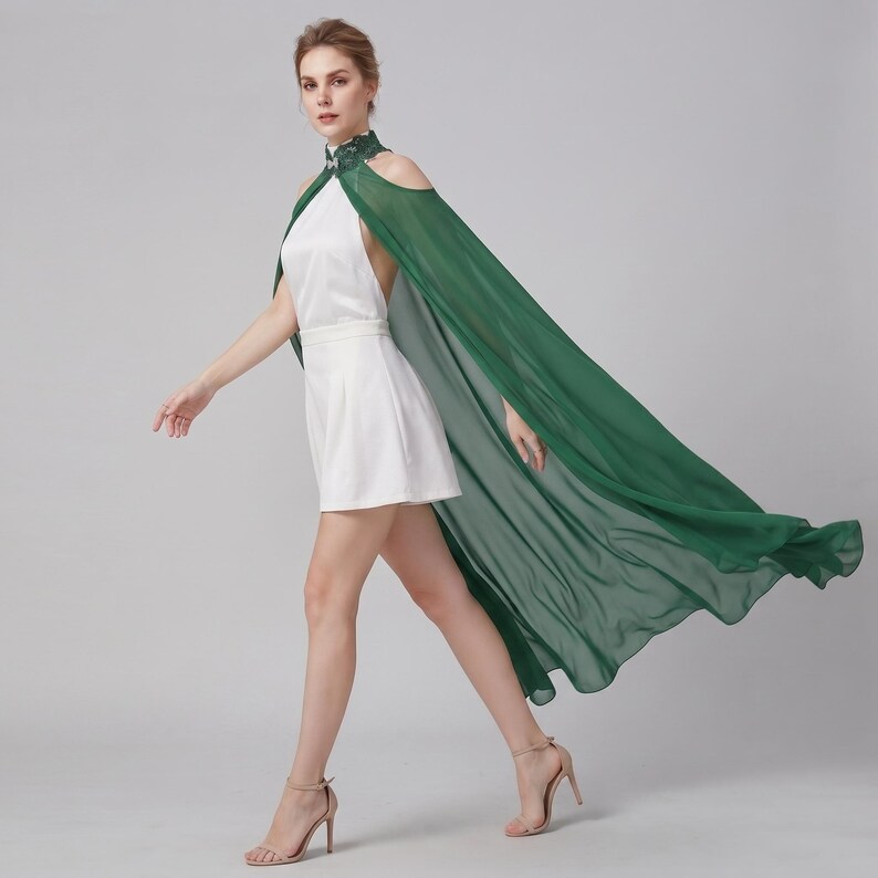 May include: A white romper featuring a high halter neckline with dark green lace detailing and cold-shoulder cutouts. Attached is a long, flowing, sheer dark green cape that extends from the shoulders to the floor, creating a dramatic silhouette.