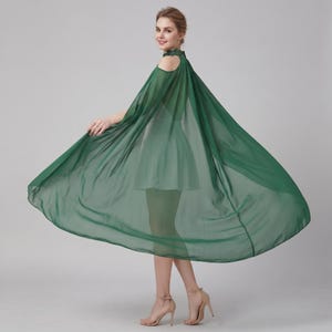 May include: A dark green dress featuring a high neckline, cold-shoulder cutouts, and a sheer, flowing cape-like overlay that extends from the shoulders. The garment has a fitted bodice and a short, flared skirt, creating a layered silhouette.
