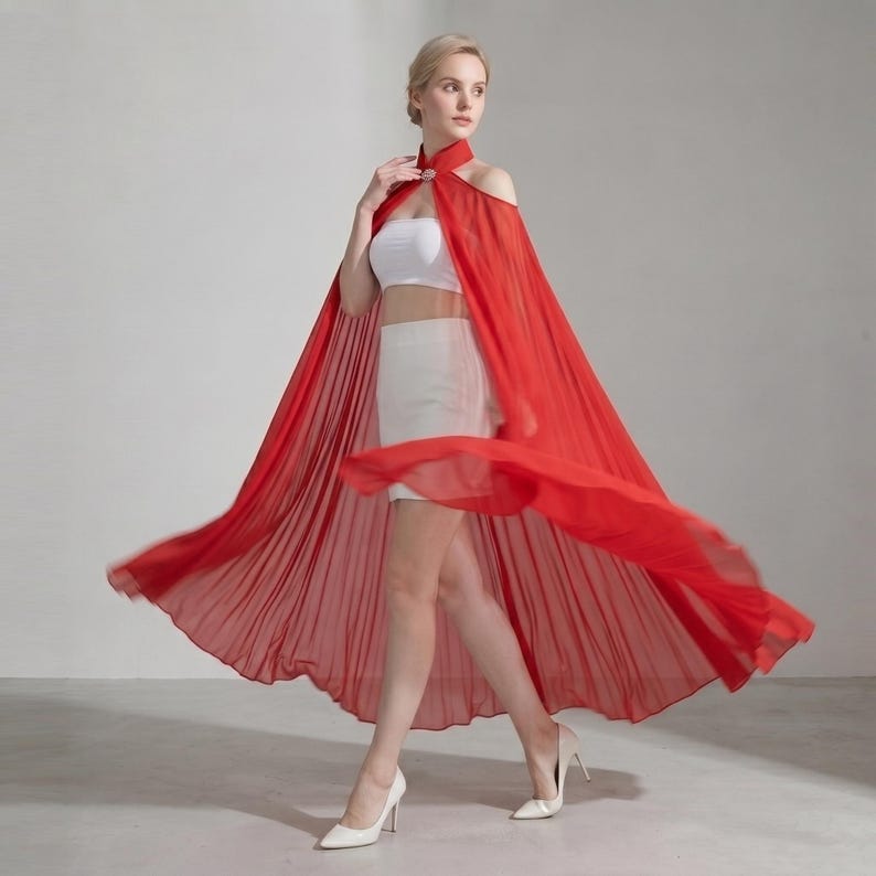 May include: A vibrant red, floor-length, pleated chiffon cape featuring a high collar with a decorative silver-toned clasp at the neck. The cape is worn over a white strapless crop top and a matching white mini skirt, paired with white heels.