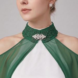 May include: A dark green high-neck collar featuring intricate floral lace embroidery and a central silver-toned metal clasp adorned with clear crystals. The collar is attached to a sheer green fabric overlay worn over a solid white garment.