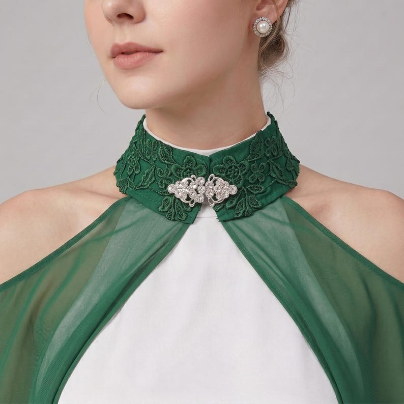 May include: A dark green high-neck collar featuring intricate floral lace embroidery and a central silver-toned metal clasp adorned with clear crystals. The collar is attached to a sheer green fabric overlay worn over a solid white garment.