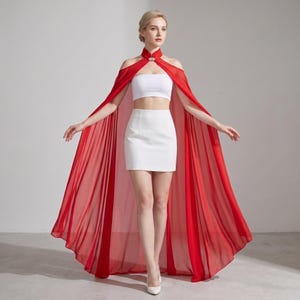 May include: A white two-piece set consisting of a strapless crop top and a high-waisted mini skirt, paired with a long, flowing red sheer cape that features a high collar and a decorative silver-toned clasp at the neckline.