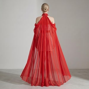 May include: A floor-length red dress featuring a high collar with intricate detailing, an open-shoulder design, and a flowing, sheer cape-like overlay that extends from the neckline to the hem, creating a layered and voluminous silhouette.