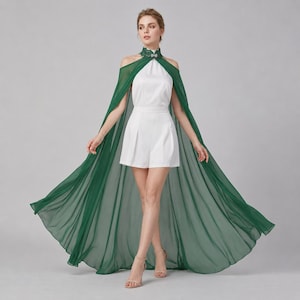 May include: A white romper featuring a high neckline with a decorative green lace collar and a flowing, floor-length sheer dark green cape attached at the shoulders. The garment has a sleeveless design with open shoulders and a tailored, pleated short bottom.