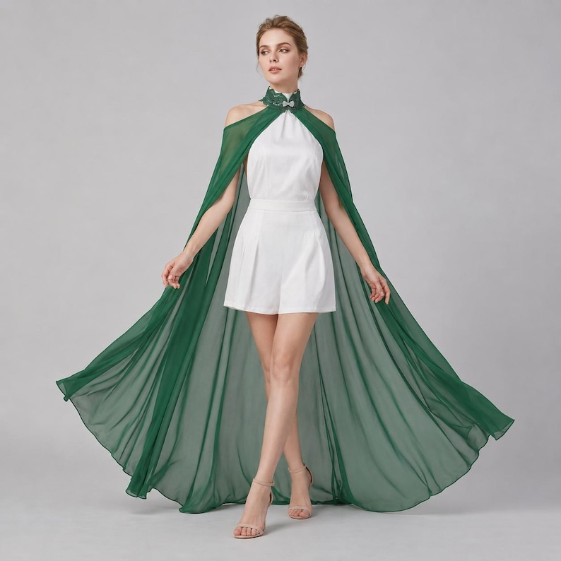 May include: A white romper featuring a high neckline with a decorative green lace collar and a flowing, floor-length sheer dark green cape attached at the shoulders. The garment has a sleeveless design with open shoulders and a tailored, pleated short bottom.