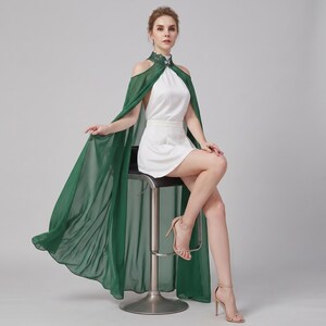 May include: A white mini dress featuring a high neck with green lace detailing and a silver embellishment. The dress includes an attached sheer dark green cape that drapes from the shoulders, creating a flowing silhouette with open shoulders.