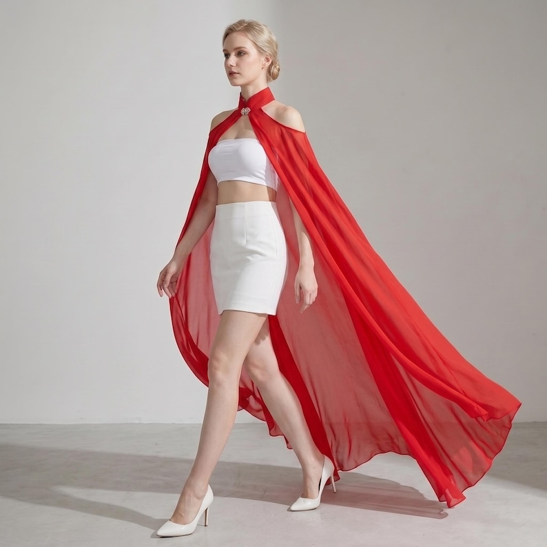 May include: A bright red floor-length chiffon cape featuring a high collar with a decorative silver clasp, worn over a white strapless crop top and a matching white mini skirt. The cape drapes from the shoulders, creating a long flowing silhouette.