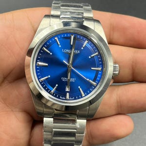 May include: A stainless steel Longines Conquest automatic wristwatch featuring a vibrant blue sunray dial, silver-tone baton hour markers, luminous hands, a date window at the six o'clock position, and a matching brushed metal link bracelet.