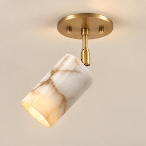 Op de afbeelding: Een plafondspot met een cilindervormige kap van wit marmer met bruine aders. Het armatuur is voorzien van een ronde voet van geborsteld messing en een bijpassende verstelbare arm die de kap met de plafondbevestiging verbindt.