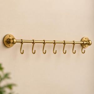 May include: A horizontal wall-mounted rail finished in a matte gold tone, featuring two circular mounting plates at each end and seven movable S-shaped hooks suspended from the central rod for hanging various items in a kitchen or entryway.