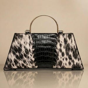 May include: A trapezoidal handbag featuring a black and white cowhide pattern on the sides and a central vertical panel of black textured material. The bag includes a single rounded gold-tone metal handle attached to the top frame.