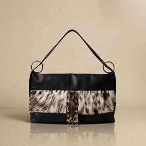May include: A black handbag featuring a horizontal panel of brown and white cowhide hair-on leather across the center. The bag includes silver-tone metal studs, a vertical leather strip, and two large silver circular rings connecting the shoulder strap.