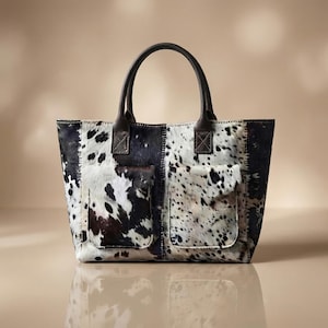 May include: A tote bag crafted from cowhide featuring a black and white spotted pattern. The bag includes two dark brown leather handles and two exterior front pockets, with visible stitching connecting the panels of the hide across the front.