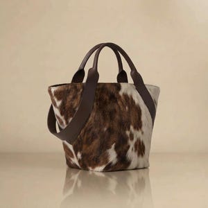May include: A tote bag featuring a brown and white cowhide pattern with a textured fur finish. The bag includes two dark brown leather handles that extend down the sides of the body, creating a structured silhouette with an open top design.