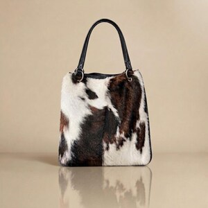 May include: A tote bag featuring a cowhide pattern with patches of white, brown, and black fur. The bag has two black leather handles attached with silver-toned metal rings at the top. The overall shape is rectangular with rounded bottom corners.