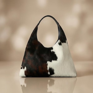May include: A hobo-style handbag featuring a cowhide pattern with patches of dark brown, black, and white fur. The bag has a triangular silhouette and a single black leather handle attached to the top, providing a structured yet soft appearance.
