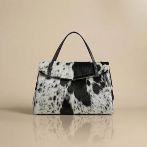 Könnte beinhalten: Eine Handtasche mit schwarz-weißem Kuhfellmuster und einer strukturierten Felloberfläche. Die Tasche verfügt über einen strukturierten Klappverschluss, einen einzelnen Tragegriff und dunkle Lederakzente an den Befestigungspunkten.