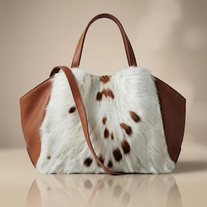 May include: A handbag featuring smooth brown leather side panels and a central panel of white faux fur with scattered brown spots. The bag includes two top handles and a long, thin brown leather shoulder strap that hangs across the front.