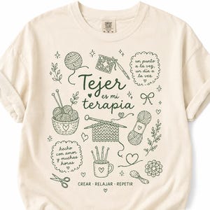 May include: A cream-colored t-shirt featuring dark green line art illustrations of knitting and crochet supplies, including yarn balls, needles, and hooks. The text reads: Tejer es mi terapia, un punto a la vez, un día a la vez, hecho con amor y muchas horas, CREAR RELAJAR REPETIR.