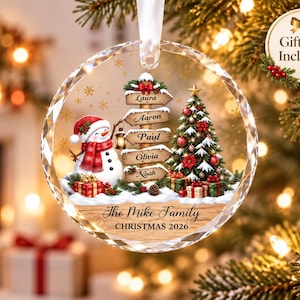 Peut inclure: Une décoration de Noël ronde en verre transparent à facettes avec une illustration imprimée d'un bonhomme de neige, d'un sapin décoré et d'un panneau en bois avec les noms Laura, Aaron, Paul, Olivia et Noah. Texte : The Mike Family CHRISTMAS 2026.