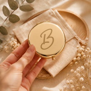 May include: A round, gold-coloured metal compact mirror featuring a large, engraved letter B in a cursive script font on the front surface. The compact has a polished, reflective finish and a small hinge mechanism visible at the top edge.