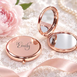 May include: Two round rose gold compact mirrors with a polished metallic finish. One mirror is closed, featuring the engraved name Emily with a small butterfly icon. The second mirror is open, revealing two reflective surfaces connected by a hinge.