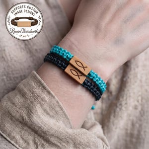 Könnte beinhalten: Zwei geflochtene Kordelarmbänder, eines in Türkis und eines in Schwarz, mit jeweils einem rechteckigen Holzelement in der Mitte, das mit einem Fischsymbol graviert ist. Das kreisförmige Logo in der Ecke zeigt den Text Supports Custom Image Designs Rowan Threadworks.